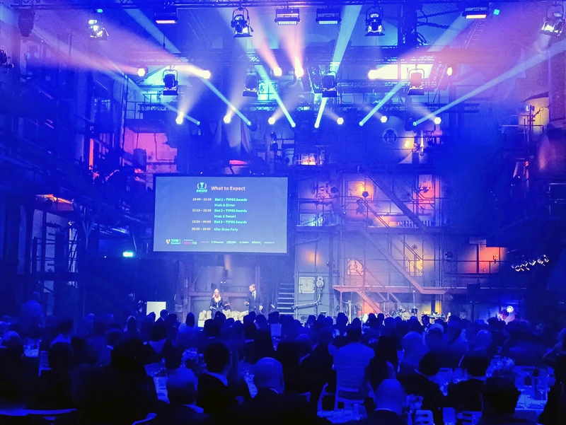 Weitwinkelaufnahme der TYPO3 Awards Gala in der historischen Alten Federnfabrik. Ein großes Publikum sitzt an runden Tischen, während die Moderatoren auf der Bühne sprechen, beleuchtet von blauem und orangefarbenem Industrielicht.