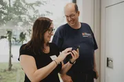 Anna Stephan zeigt Jens Krumm etwas auf dem Smartphone. Beide lächeln und stehen in einem hellen Raum mit Blick ins Grüne. Die Szene wirkt gelöst und kollegial.