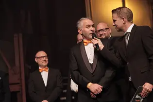 Matthias Hinner von +Pluswerk Earlybird spricht auf der Bühne der TYPO3 Award Gala 2025 in ein Mikrofon, während neben ihm weitere Teammitglieder stehen und lächeln.