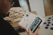 Eine Person hält ein Smartphone und ein Blatt Papier mit Blindenschrift (Brailleschrift) in der Hand. Im Hintergrund liegen Stoffbeutel und Spielkarten auf einem Tisch. Szene aus dem Barrierefreiheits-Workshop beim +Pluscamp 2025.