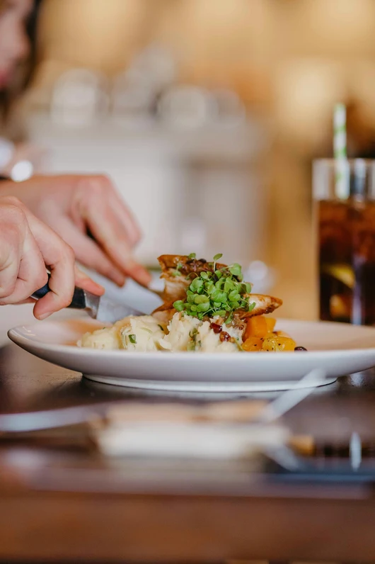 Nahaufnahme eines angerichteten warmen Gerichts auf einem Teller in einem Restaurant. Eine Person schneidet das Essen mit Messer und Gabel. Im Hintergrund steht ein Getränk auf dem Tisch.