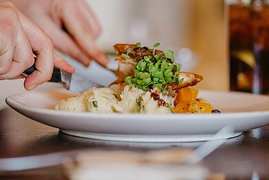 Nahaufnahme eines angerichteten warmen Gerichts auf einem Teller in einem Restaurant. Eine Person schneidet das Essen mit Messer und Gabel. Im Hintergrund steht ein Getränk auf dem Tisch.