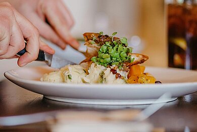 Close-up of a hot dish on a plate in a restaurant. A person cuts the food with a knife and fork. A drink is on the table in the background.