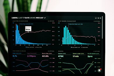 DE Ein Laptopbildschirm zeigt eine Datenanalyse mit mehreren Diagrammen und Metriken zu Nutzerverhalten, Ladezeiten und Absprungraten. Die Benutzerstatistiken der letzten sieben Tage sind visualisiert, mit blauen Balken und pinken Linien, die Trends darstellen. Im Hintergrund sind unscharfe grüne Blätter sichtbar. EN A laptop screen shows a data analysis with several diagrams and metrics on user behaviour, loading times and bounce rates. The user statistics of the last seven days are visualised, with blue bars and pink lines representing trends. Blurred green leaves are visible in the background.