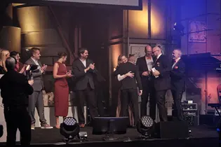 Auf der Bühne der TYPO3 Award Gala 2025 umarmen sich Teammitglieder von +Pluswerk und Earlybird nach der Preisverkündung. Andere Personen auf der Bühne applaudieren, während ein Kameramann den Moment festhält.