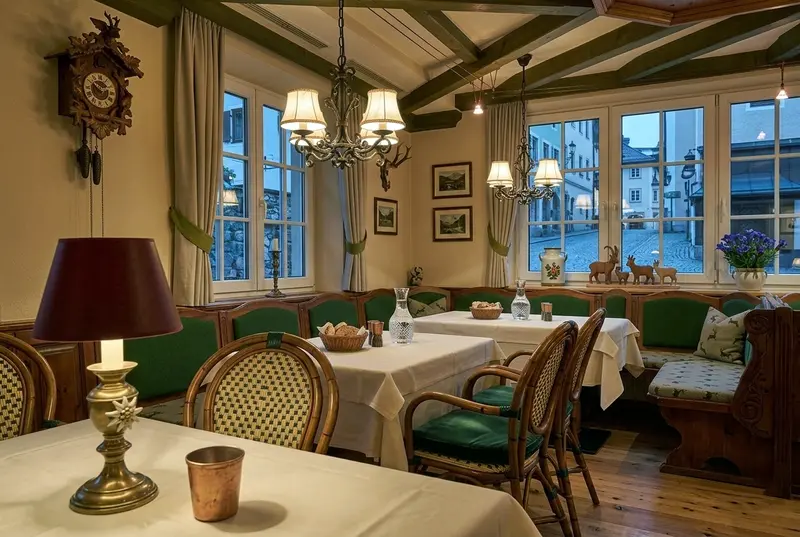 Gemütlicher Gastraum eines traditionellen Restaurants mit Holzbalkendecke, Kronleuchtern und mehreren gedeckten Tischen mit weißen Tischdecken. Entlang der Fenster stehen grün gepolsterte Sitzbänke und Holzstühle. An den Wänden hängen Bilder und eine Kuckucksuhr.