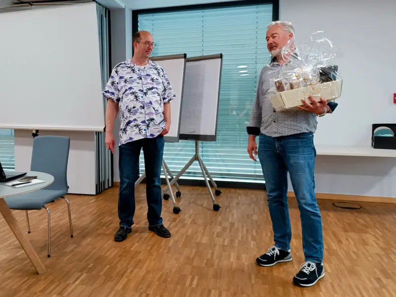 Jens Krumm (links) und Hans-Jörg Raphael (rechts) während einer Präsentation beim +Pluscamp 2025 in Rüsselsheim; Hans-Jörg hält einen Geschenkkorb, im Hintergrund sind Logos der +Pluswerk-Partner auf einer Leinwand zu sehen.