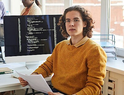 A young software developer wearing glasses and a mustard-colored sweater sits at a desk in a modern office, holding documents. Behind him, a computer screen displays lines of code. In the background, three colleagues collaborate, working on laptops and tablets in a bright, open workspace.