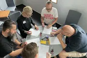 Sechs Personen sitzen um einen Tisch und bearbeiten konzentriert Arbeitsblätter mit Brailleschrift. In der Mitte liegen Stifte und Hilfsmittel. 