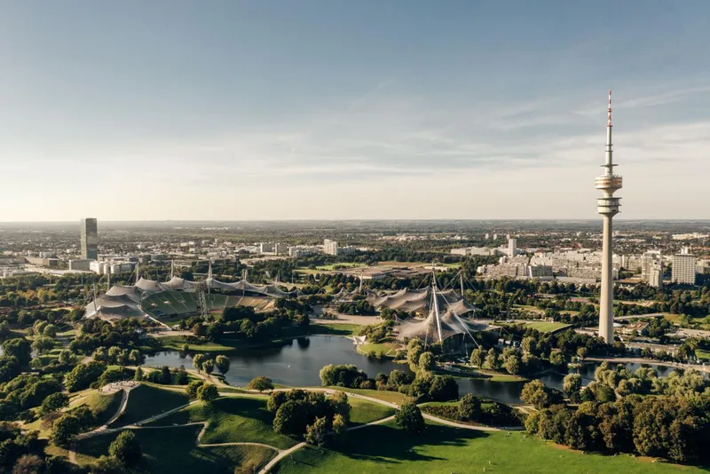 Olympiapark München – Luftaufnahme mit Olympiaturm Luftaufnahme des Olympiaparks München mit Olympiaturm, Zeltdach und Seen bei klarem Himmel.