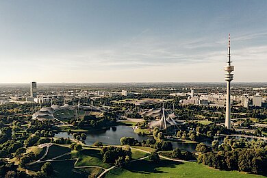 Luftaufnahme des Olympiaparks München mit Olympiaturm, Zeltdach und Seen bei klarem Himmel.