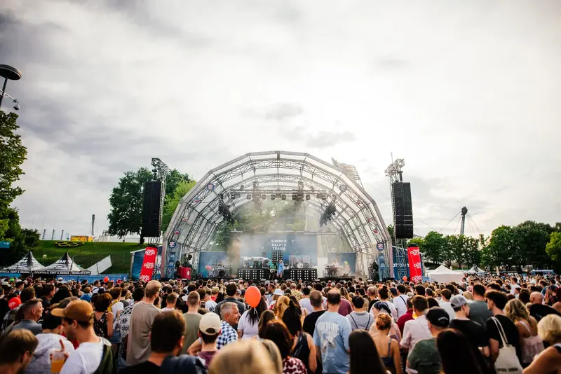 Sommernachtstraum im Olympiapark München mit Konzertbühne und Festivalpublikum Besucherinnen und Besucher verfolgen ein Live-Konzert auf der Hauptbühne des Sommernachtstraums im Olympiapark München. Das Bild zeigt die Festivalatmosphäre mit großer Bühne, dichtem Publikum und sommerlichem Abendhimmel.