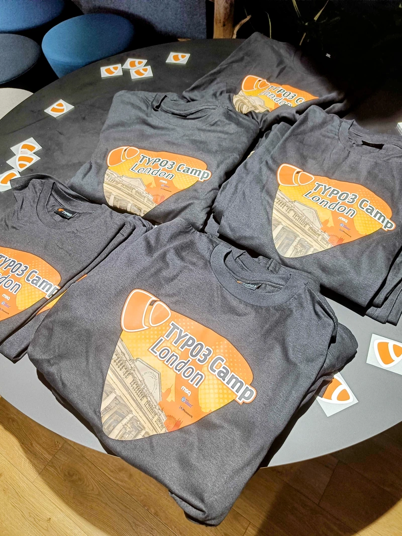 TYPO3 Camp London T-Shirts für die Community Mehrere TYPO3 Camp London T-Shirts mit orangefarbenem Logo und Aufdruck liegen auf einem runden Tisch bereit für die Teilnehmenden.