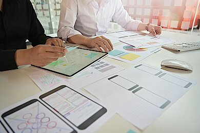 Zwei Personen arbeiten an UX- und UI-Designs mit Tablet und ausgedruckten Wireframes.