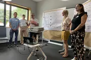 Fünf Personen stehen in einem Workshopraum vor zwei Flipcharts mit großformatigen Ausdrucksbögen. Eine Person spricht, die anderen hören zu.