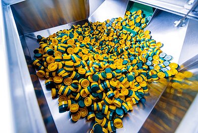 Große Menge gelb-grüner Kunststoffverschlüsse in einer industriellen Produktionsanlage für PET-Verpackungen, unsortiert in einem Metallbehälter.