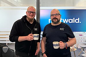 Zwei Männer stehen in einer Büroumgebung mit gezückten Kaffeetassen vor einer halbdurchsichtigen Glasschiebetür. Links steht Hannes Strangmeier, rechts Martin Helmich.