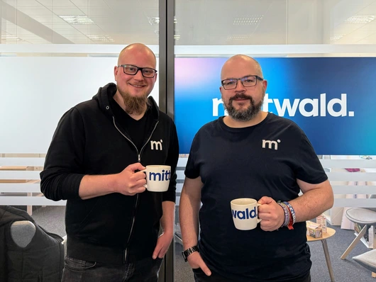 Zwei Männer stehen in einer Büroumgebung mit gezückten Kaffeetassen vor einer halbdurchsichtigen Glasschiebetür. Links steht Hannes Strangmeier, rechts Martin Helmich.