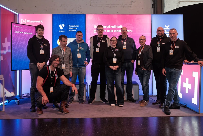 +Pluswerk and Incluthon team at the T3CON25 trade fair stand Gruppenfoto des +Pluswerk Teams am Messestand der T3CON25. Elf Personen stehen und knien vor den farbig beleuchteten TYPO3- und +Pluswerk-Hintergründen und blicken lächelnd in die Kamera.