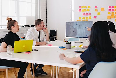 Team sitting at table and analysing together with computer display and Kanban notes at the wall.