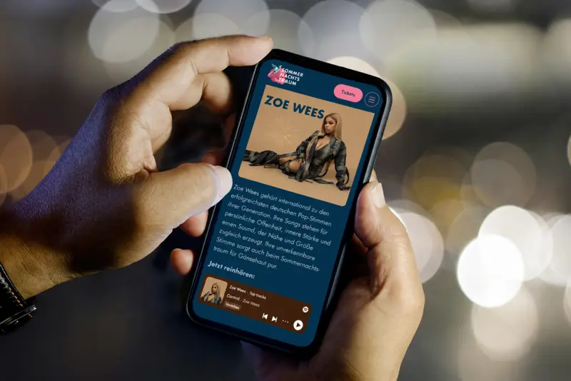 Mobile Ansicht der Sommernachtstraum-Website auf einem Smartphone Ein Smartphone zeigt die mobile Ansicht der Sommernachtstraum-Website. Zu sehen sind die Künstlerseite von Zoe Wees, Informationen zum Auftritt sowie ein integrierter Musik-Player. Die Website ist für die mobile Nutzung optimiert und bietet einen direkten Zugang zu Programm und Tickets.