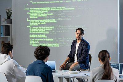 Ein Referent erklärt vor einer projizierten Codeansicht die Struktur einer Website, während drei Teilnehmende an einem Tisch mit Laptops zuhören.