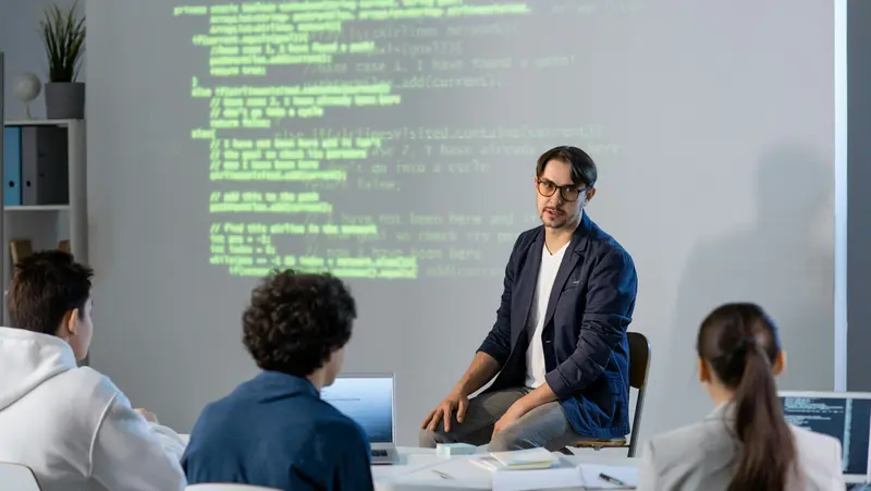 KI-Workshop zur Analyse von Website-Struktur und Code Ein Referent erklärt vor einer projizierten Codeansicht die Struktur einer Website, während drei Teilnehmende an einem Tisch mit Laptops zuhören.