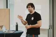 Stefan Barac steht an einem Stehtisch mit Laptop und spricht während seines Workshops beim +Pluscamp 2025. Er gestikuliert leicht und trägt ein schwarzes T-Shirt.