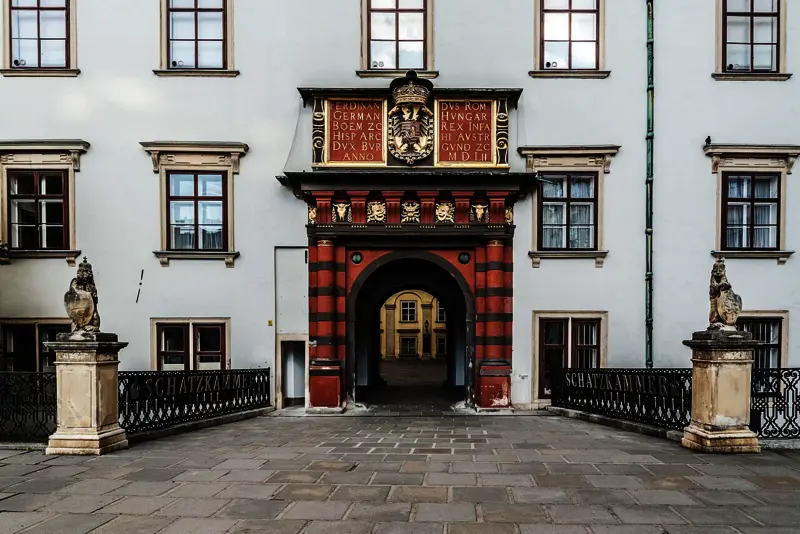 Historisches Tor der Wiener Hofburg, das sogenannte Schweizertor, mit roter und schwarzer Steinverzierung sowie goldenen Wappen- und Inschriftenfeldern; Zugang zur Schatzkammer des Bundesdenkmalamts.
