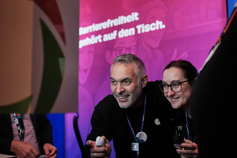 Matthias Hinner and Anna Stephan at the photo box at the +Pluswerk stand Matthias Hinner und Anna Stephan von +Pluswerk posieren lachend an der Fotobox des Messestands. Im Hintergrund ist ein pink beleuchtetes Banner mit der Aufschrift „Barrierefreiheit gehört auf den Tisch“ zu sehen.