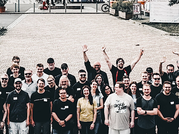 Die Mitarbeiter von +Pluswerk stehen im Freien am Main-Ufer in Rüsselsheim, winken fröhlich in die Kamera und grüßen zu einem Gruppenfoto.