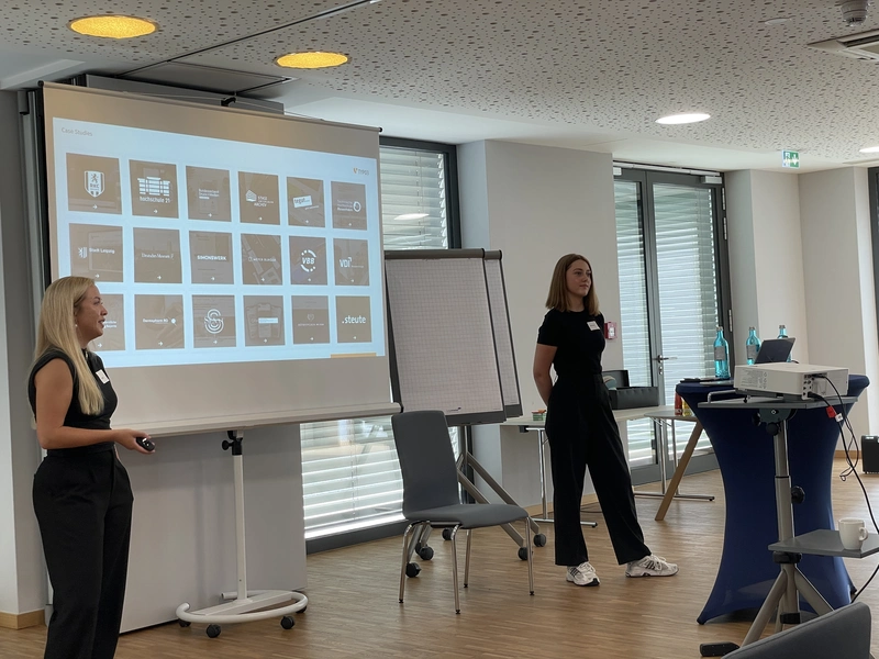 Zwei Referentinnen stehen neben einer Leinwand und präsentieren vor Publikum. Auf der Folie sind viele kleine Icons oder Screenshots zu sehen. Die Szene zeigt die Session ‚TYPO3 im Wandel‘ von Luisa Faßbender und Myrna Gönnemann.