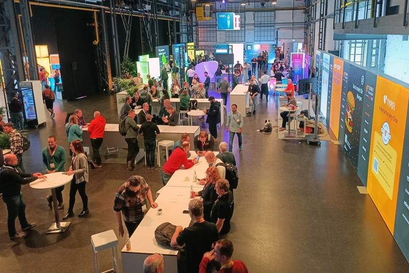 T3CON25 Networking and exhibitor area in the Areal Böhler Blick in die Networking- und Ausstellerhalle der T3CON25: Besucher stehen und sitzen an hohen Tischen, führen Gespräche und erkunden die Stände unterschiedlicher Aussteller.