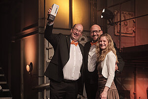 Drei Mitglieder des Earlybird- und +Pluswerk-Teams posieren auf der Bühne mit der TYPO3-Auszeichnung in den Händen und strahlen vor dem Hintergrund der industriellen Beleuchtung.