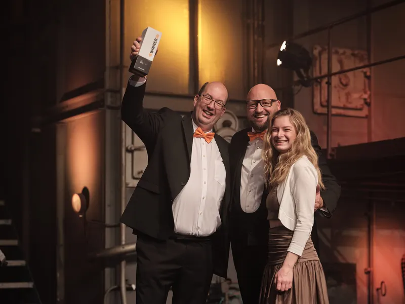 Drei Mitglieder des Earlybird- und +Pluswerk-Teams posieren auf der Bühne mit der TYPO3-Auszeichnung in den Händen und strahlen vor dem Hintergrund der industriellen Beleuchtung.