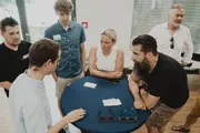 Sechs Personen stehen um einen runden Stehtisch mit blauer Tischdecke und diskutieren aktiv. Auf dem Tisch liegen Spielkarten und andere Workshop-Materialien.