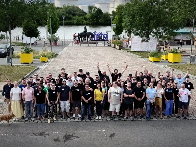 Gruppenfoto von etwa 60 Teilnehmern des Pluscamps 2025, aufgenommen im Freien auf einem gepflasterten Platz in Rüsselsheim mit Blick auf den Main. Die Personen stehen dicht beieinander, einige heben die Arme jubelnd in die Luft. Im Hintergrund ist ein Reiterstandbild, ein „RÜSSELSHEIM“-Schriftzug und viel Grün zu sehen. Ein Hund steht links am Bildrand.