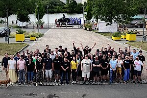 Gruppenfoto von etwa 60 Teilnehmern des Pluscamps 2025, aufgenommen im Freien auf einem gepflasterten Platz in Rüsselsheim mit Blick auf den Main. Die Personen stehen dicht beieinander, einige heben die Arme jubelnd in die Luft. Im Hintergrund ist ein Reiterstandbild, ein „RÜSSELSHEIM“-Schriftzug und viel Grün zu sehen. Ein Hund steht links am Bildrand.