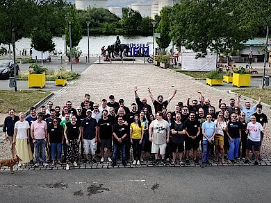 Gruppenfoto von etwa 60 Teilnehmern des Pluscamps 2025, aufgenommen im Freien auf einem gepflasterten Platz in Rüsselsheim mit Blick auf den Main. Die Personen stehen dicht beieinander, einige heben die Arme jubelnd in die Luft. Im Hintergrund ist ein Reiterstandbild, ein „RÜSSELSHEIM“-Schriftzug und viel Grün zu sehen. Ein Hund steht links am Bildrand.