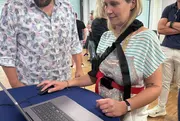 Eine Frau trägt eine Tremor-Simulationsweste und versucht, mit zitternden Bewegungen eine Aufgabe am Laptop zu lösen. Ein Mann steht neben ihr und beobachtet interessiert.