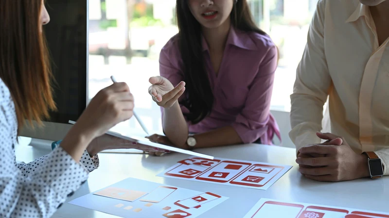 UX/UI design and digital user experiences Three people are sitting around a conference table discussing designs for a digital user interface and mobile applications.
