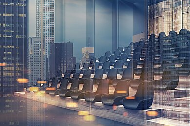 Leerer, moderner Vortragssaal mit mehreren Reihen dunkler Sitze, aufgenommen in Seitenansicht. Durch die Glasfront spiegelt sich eine urbane Skyline aus hohen Bürogebäuden. Warmes Streulicht erzeugt eine leichte Unschärfe und Atmosphäre von Vorbereitung vor einer Veranstaltung.