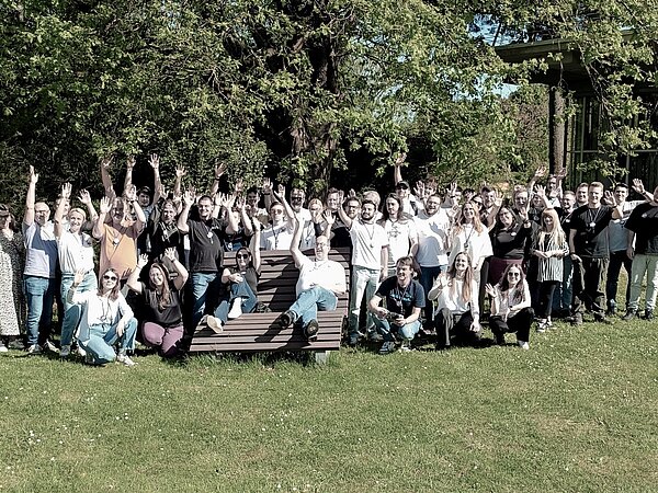 Die +Pluswerk Crew in Karlsruhe beim +Pluscamp Die Mitarbeiter von +Pluswerk stehen im Grünen in Karlsruhe, winken fröhlich in die Kamera und grüßen zu einem Gruppenfoto.