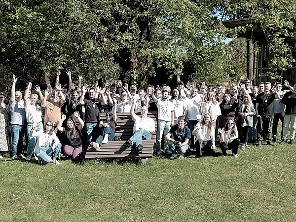 Die Mitarbeiter von +Pluswerk stehen im Grünen in Karlsruhe, winken fröhlich in die Kamera und grüßen zu einem Gruppenfoto.