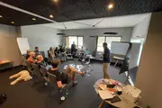 Mehrere Personen sitzen im Kreis in einem Workshopraum. Eine Person steht vorne und spricht, Flipcharts und Präsentationsmaterialien sind im Raum verteilt.