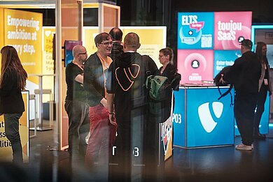 People interacting and networking at the TYPO3 booth area during T3CON25, surrounded by illuminated displays and colorful brand panels.